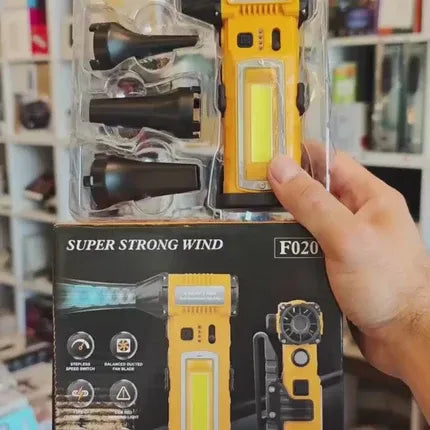 A hand holding a packaged yellow and black portable flashlight with attached accessories in a clear plastic blister pack, with the retail box below it. The box reads 'SUPER STRONG WIND' and shows the model 'F020'.
