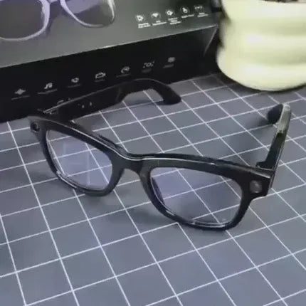 Black rectangular eyeglasses with clear lenses resting on a gray grid cutting mat, with a black product box and a pale green ceramic pot in the background. Bluetooth Smart Glasses for Travel and Daily Use UAE peetta